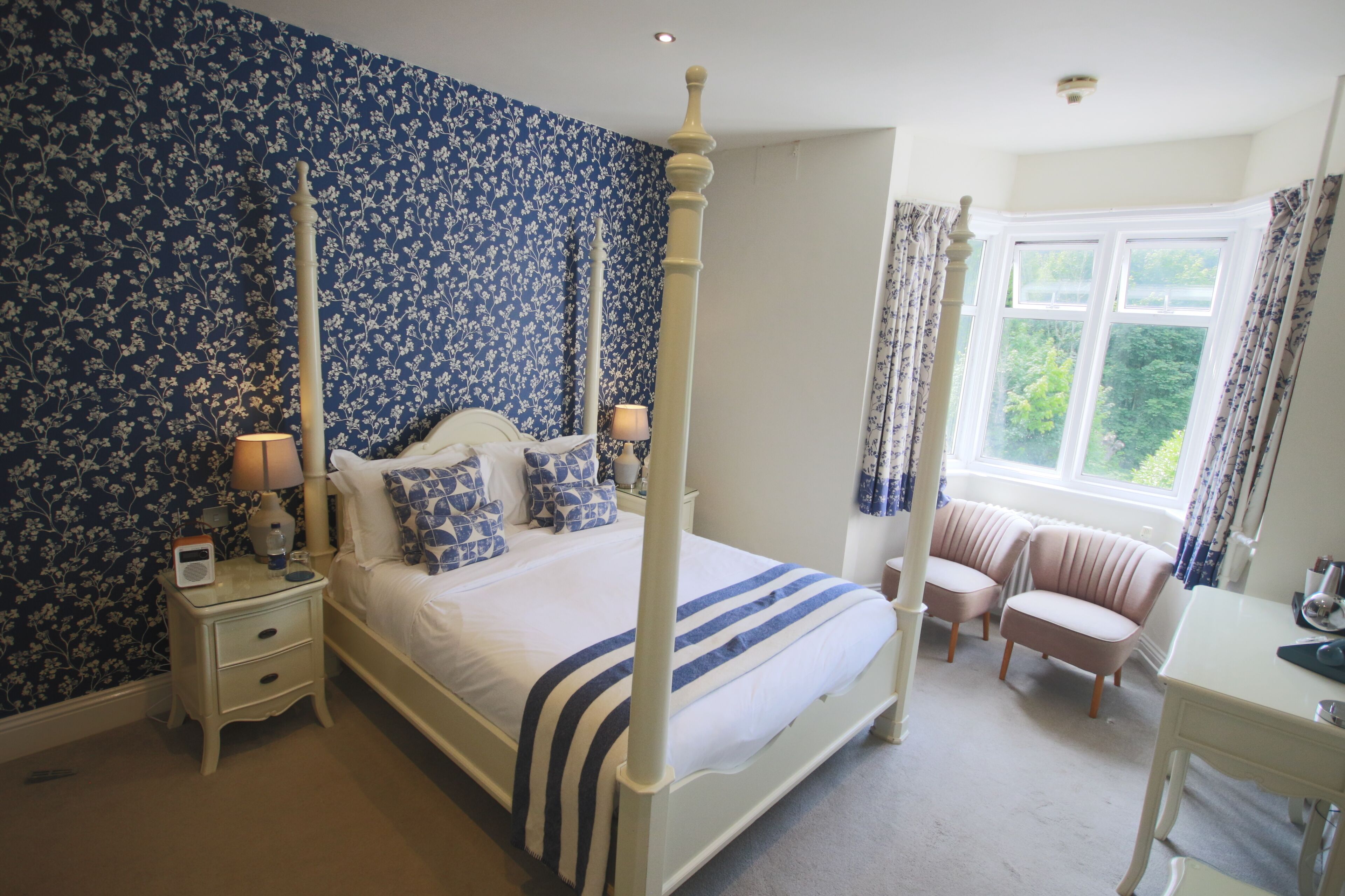 A four-poster bed with a white and blue striped bedspread, flanked by two bedside tables with lamps, a nightstand, and a window with curtains.