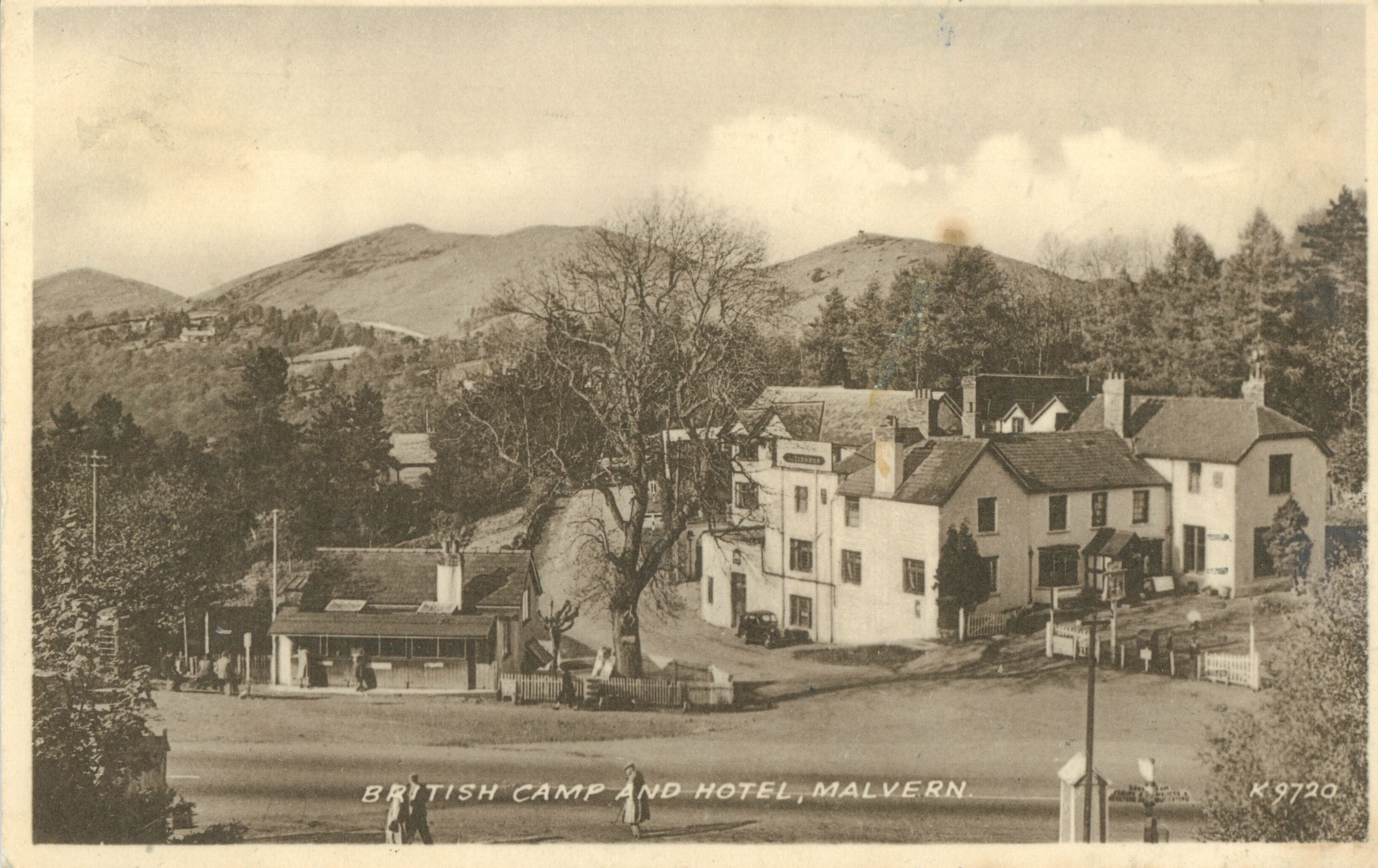 A British Camp and Hotel in Malvern with a prominent building, a small structure with a sign, and a hill in the background.
