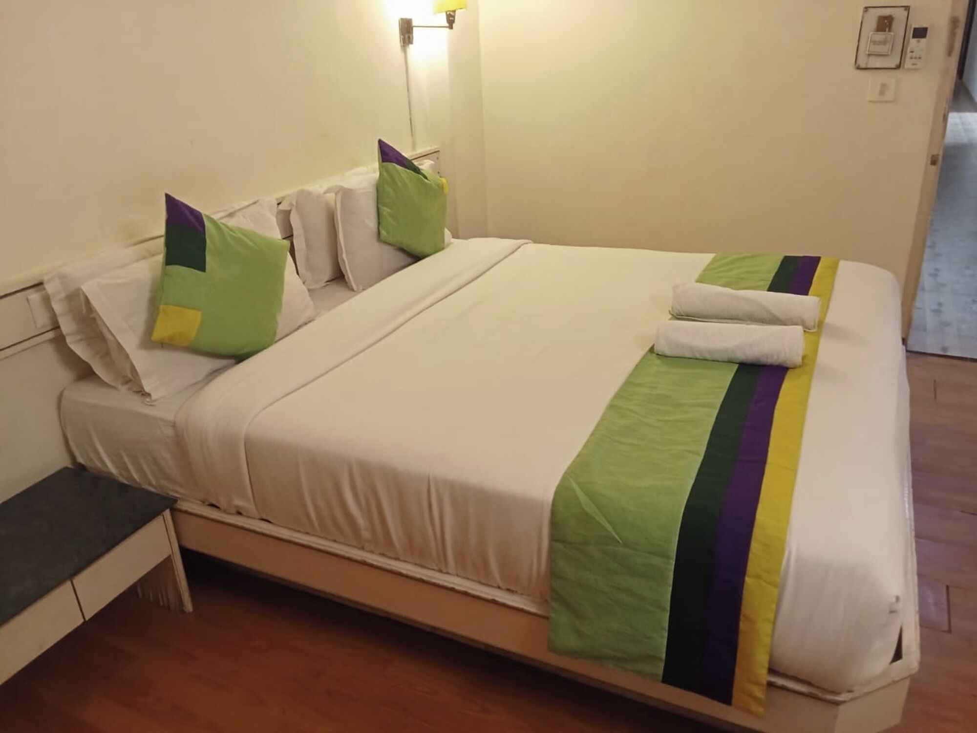 Una cama bien hecha con una manta verde y morada a rayas, almohadas blancas y una mesita de noche con una lámpara.