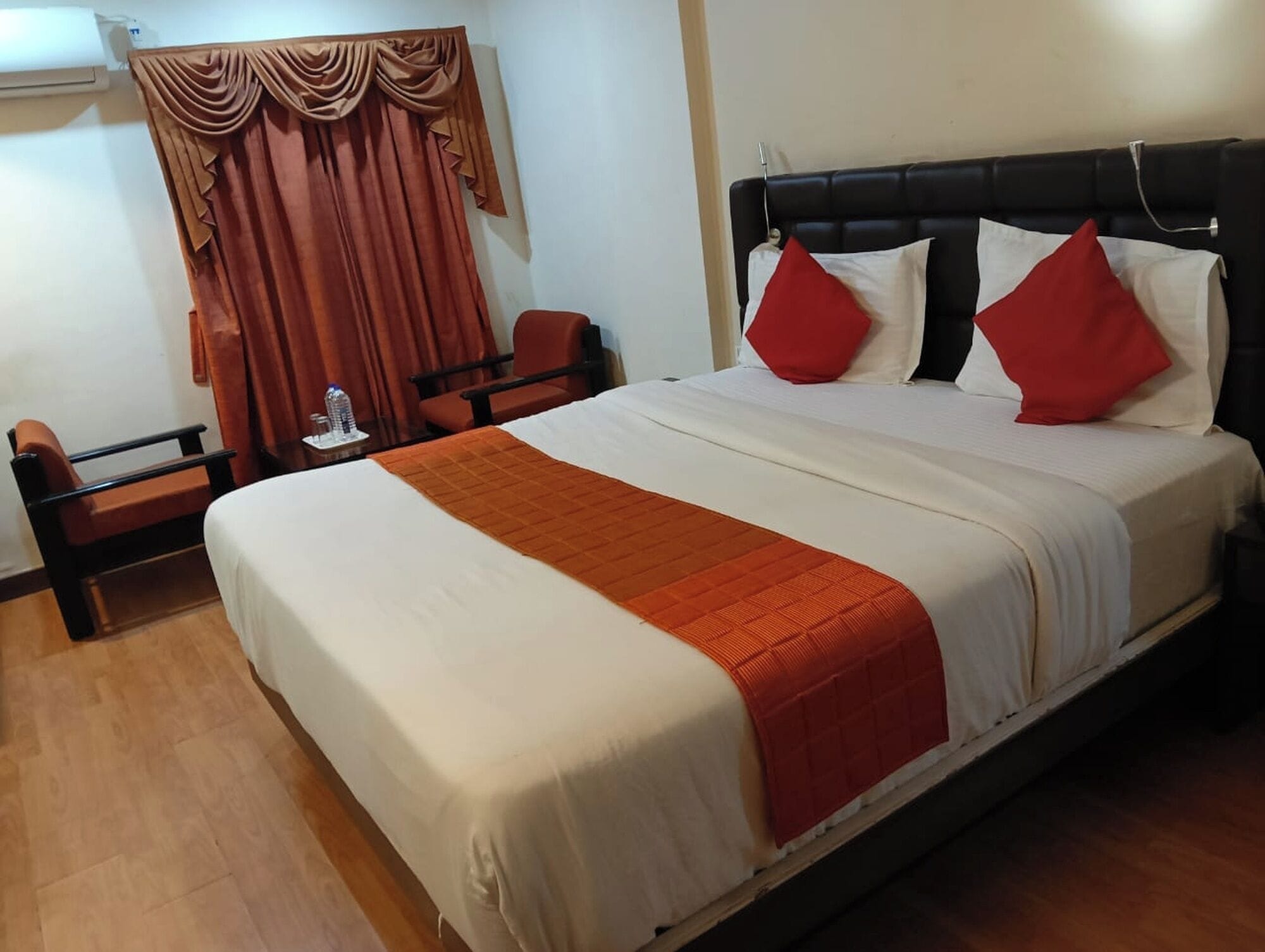 Habitación de hotel con cama, dos sillas, una mesa con una botella y un ventanal con cortinas.