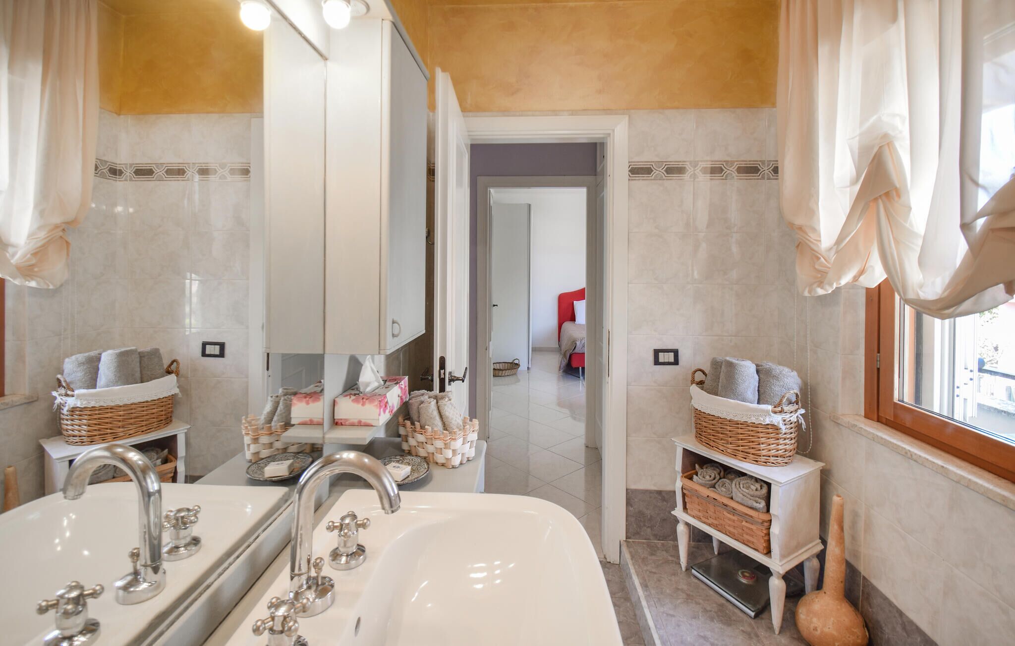 Une salle de bain dotée d’un grand miroir, de deux lavabos et d’une douche. Des paniers en osier contenant des serviettes et des articles de toilette sont disposés sur des étagères.
