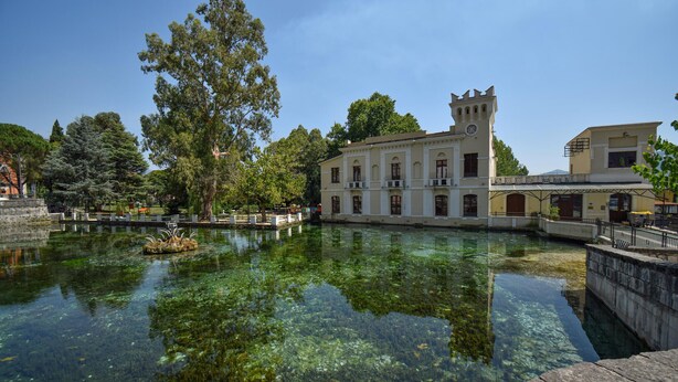 Un edificio histórico con una torre, un estanque tranquilo que refleja la estructura y árboles circundantes.