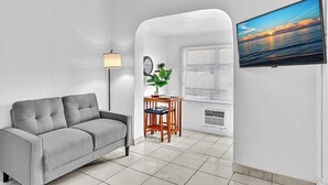Un salon moderne avec un canapé gris, une petite table d’appoint en bois sur laquelle est posée une plante, un téléviseur fixé au mur et une fenêtre avec des stores.