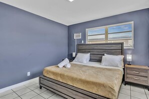 A bedroom with a bed, nightstands, a window with blinds, and a thermostat on the wall.