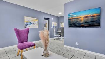 A hotel lobby with a purple chair, a white table, a vase with pampas grass, and a TV displaying a sunset view.