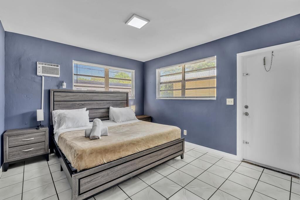 Une chambre à coucher comprenant un lit, des tables de chevet, un climatiseur fixé au mur et des fenêtres équipées de stores.