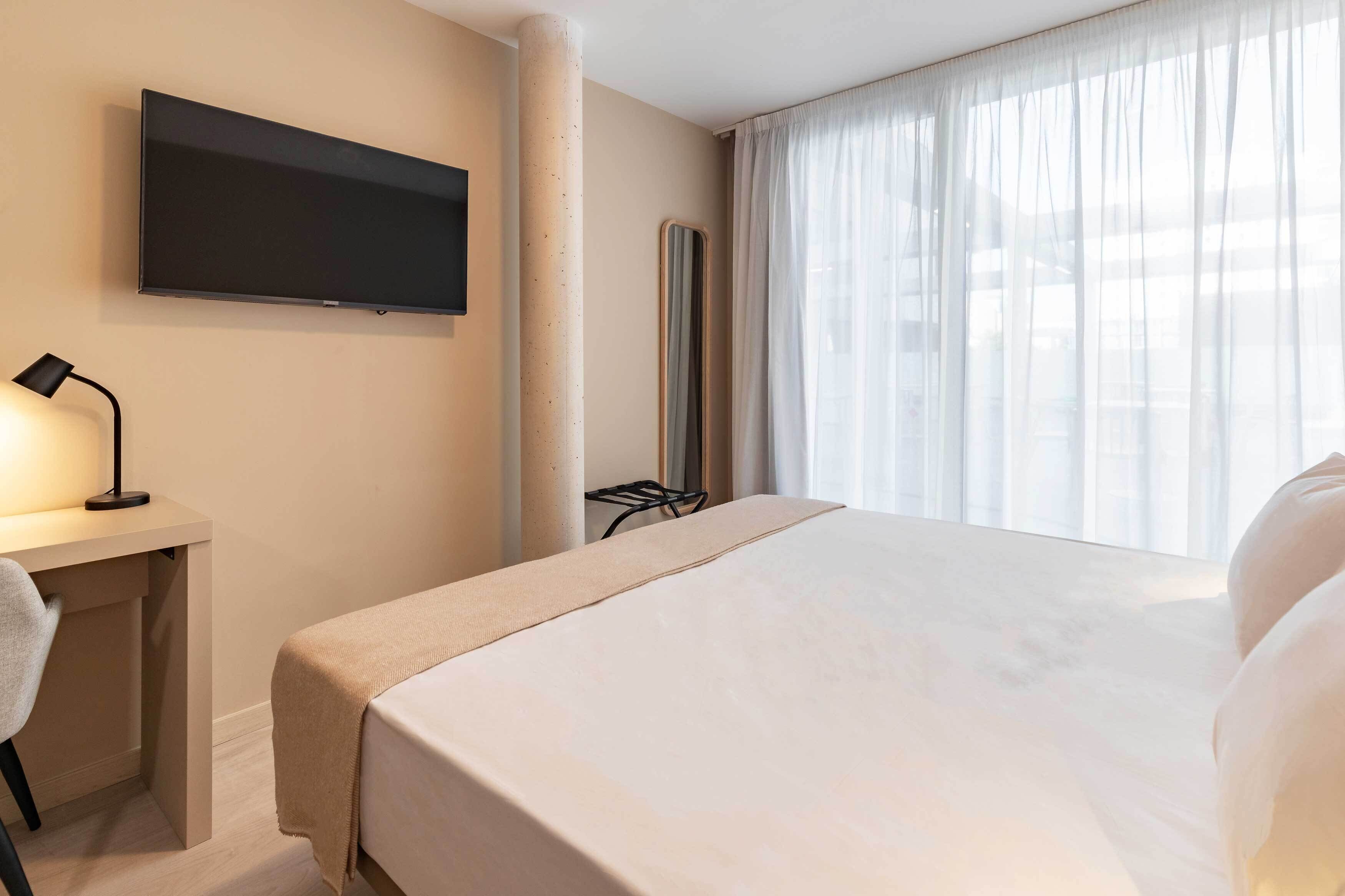 Habitación de hotel con cama, escritorio con silla, televisor montado en la pared y ventana con cortinas traslúcidas.