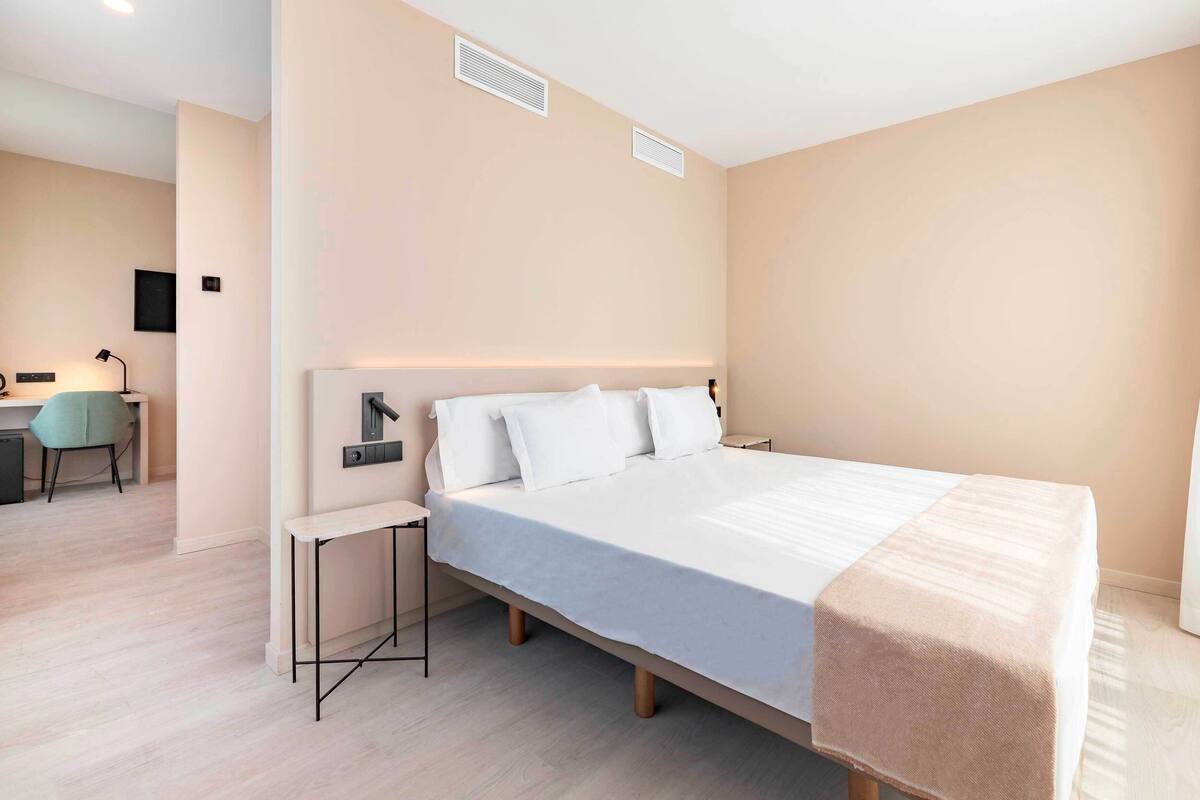 A modern hotel room with a large bed, bedside tables, a desk with a chair, and a small desk lamp.