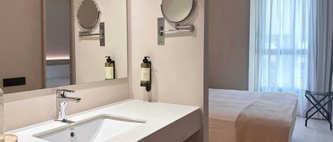 Un baño de hotel con un lavamanos grande, dos espejos adheridos a la pared y una cama con una colcha blanca.