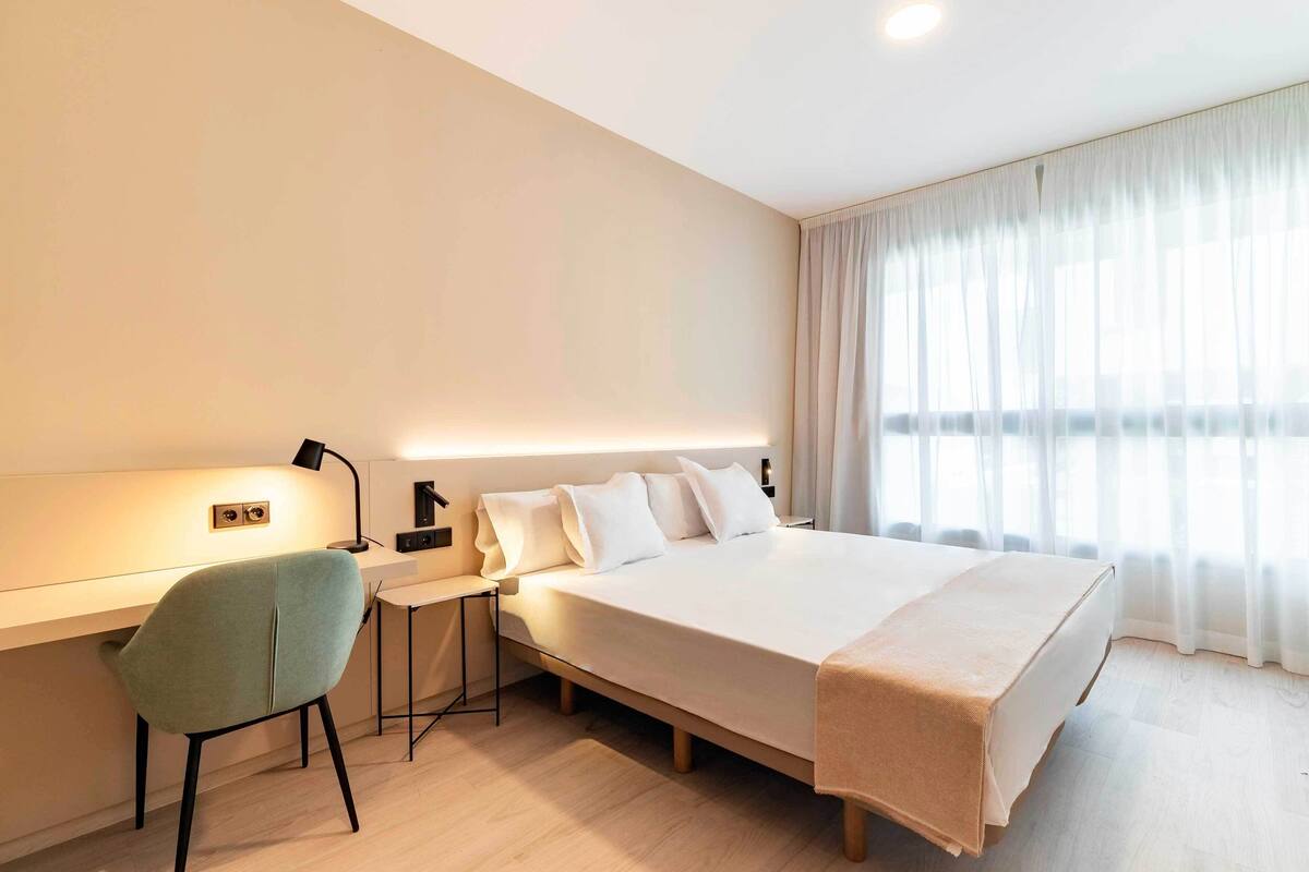 A modern hotel room with a large bed, a desk with a chair, and a bedside table.