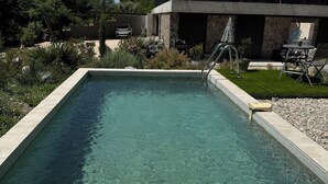 A rectangular swimming pool with clear water, surrounded by a pebble border, a stone wall, and a modern building with a balcony. - Two Gîtes With Swimming Pool les Rochers de Plaisians (PLAISIANS)