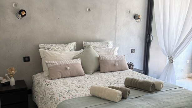 Een moderne slaapkamer met een betonnen muur, een bed met bloemenbedrukking, een nachtkastje met een vaas en een groot raam met dunne gordijnen.