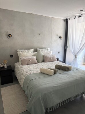 A modern bedroom with a concrete wall, a bed with floral bedding, a bedside table with a vase, and a large window with sheer curtains.