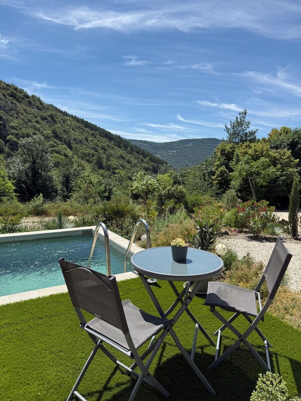 A pool with a view of a mountainous landscape, surrounded by greenery and a small table with chairs.