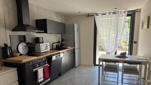Een kleine keuken met moderne apparatuur, een eethoek met een tafel en stoelen, en uitzicht door een raam met dunne gordijnen.