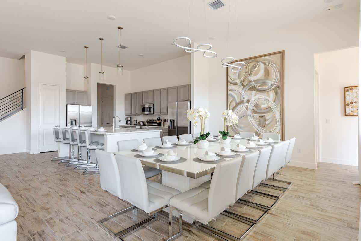 A modern kitchen with stainless steel appliances, a dining area with white chairs and a long table set for a meal, and a staircase leading to another level.