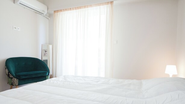 A hotel room with a large bed, a green armchair, a bedside table with a lamp, and a window with sheer curtains. - Modern 1 Bd Balcony Apt in Kato Patisia (Athens)