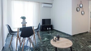 A modern living room with a glass dining table, black chairs, a small round table with a candle and a decorative item, a flat-screen TV on a black stand, and a wall-mounted air conditioner. - Modern 1 Bd Balcony Apt in Kato Patisia (Athens)