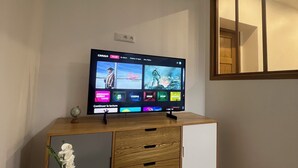 Une pièce comprenant un bureau en bois, un téléviseur affichant l’interface d’un service de diffusion en continu, une petite table blanche ornée d’un objet décoratif et un grand miroir au mur.