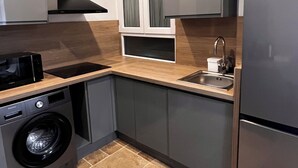 Une cuisine compacte avec des meubles gris, un réfrigérateur en acier inoxydable, un lave-linge encastré et une fenêtre au-dessus de l’évier.