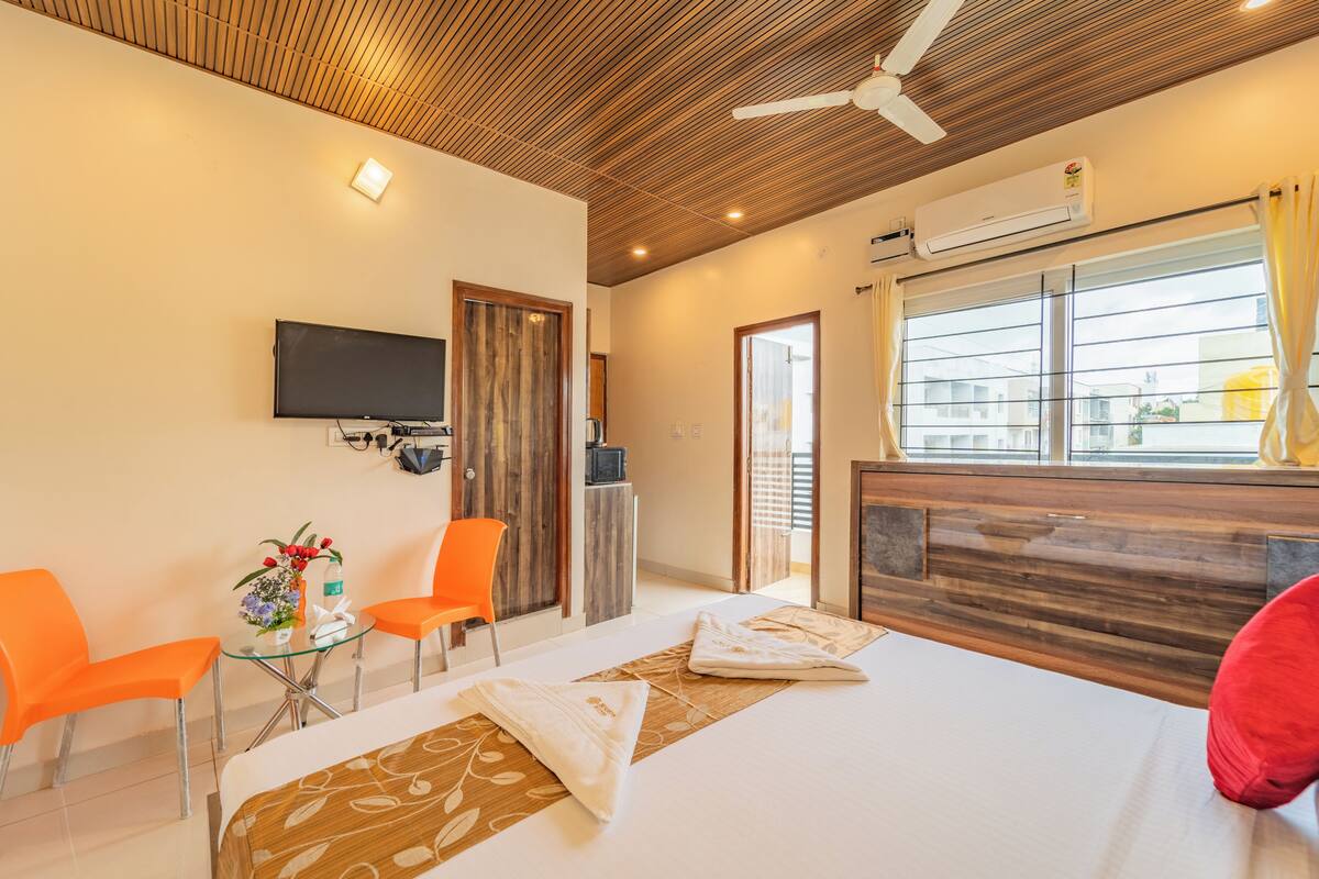 A hotel room with a bed, two orange chairs, a small table with a vase, a TV mounted on the wall, and an air conditioning unit.