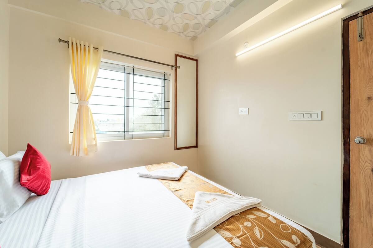 A bedroom with a bed, a window with curtains, a wooden door, and a light switch on the wall.