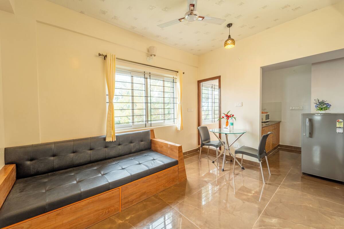 A modern living room with a sofa, a dining table with chairs, a kitchen area with a refrigerator, and large windows with curtains.