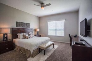 Une chambre à coucher comprenant un lit, des tables de chevet, un bureau, une chaise, un téléviseur et une fenêtre avec des stores.