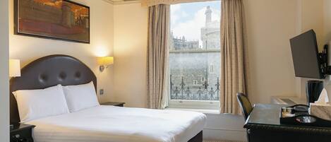 A hotel room with a large bed, a desk with a computer, a TV, and a window with a view of a historic building.