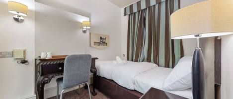 A hotel room with a bed, a desk with a chair, a lamp, and a wall-mounted shelf with a telephone.