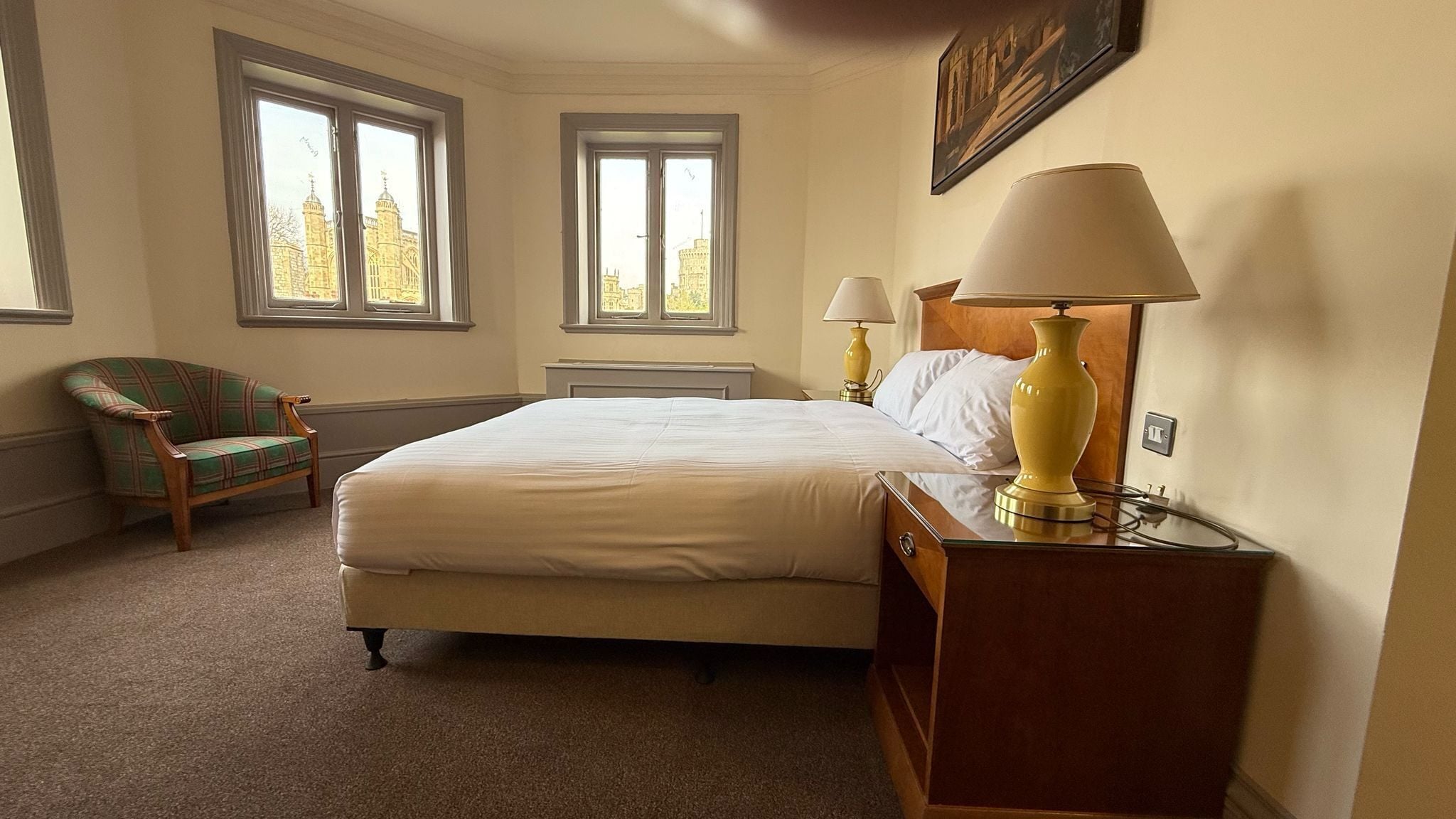 Habitación de hotel con una cama grande, dos lámparas de noche, una silla y dos ventanas con vista a la ciudad.
