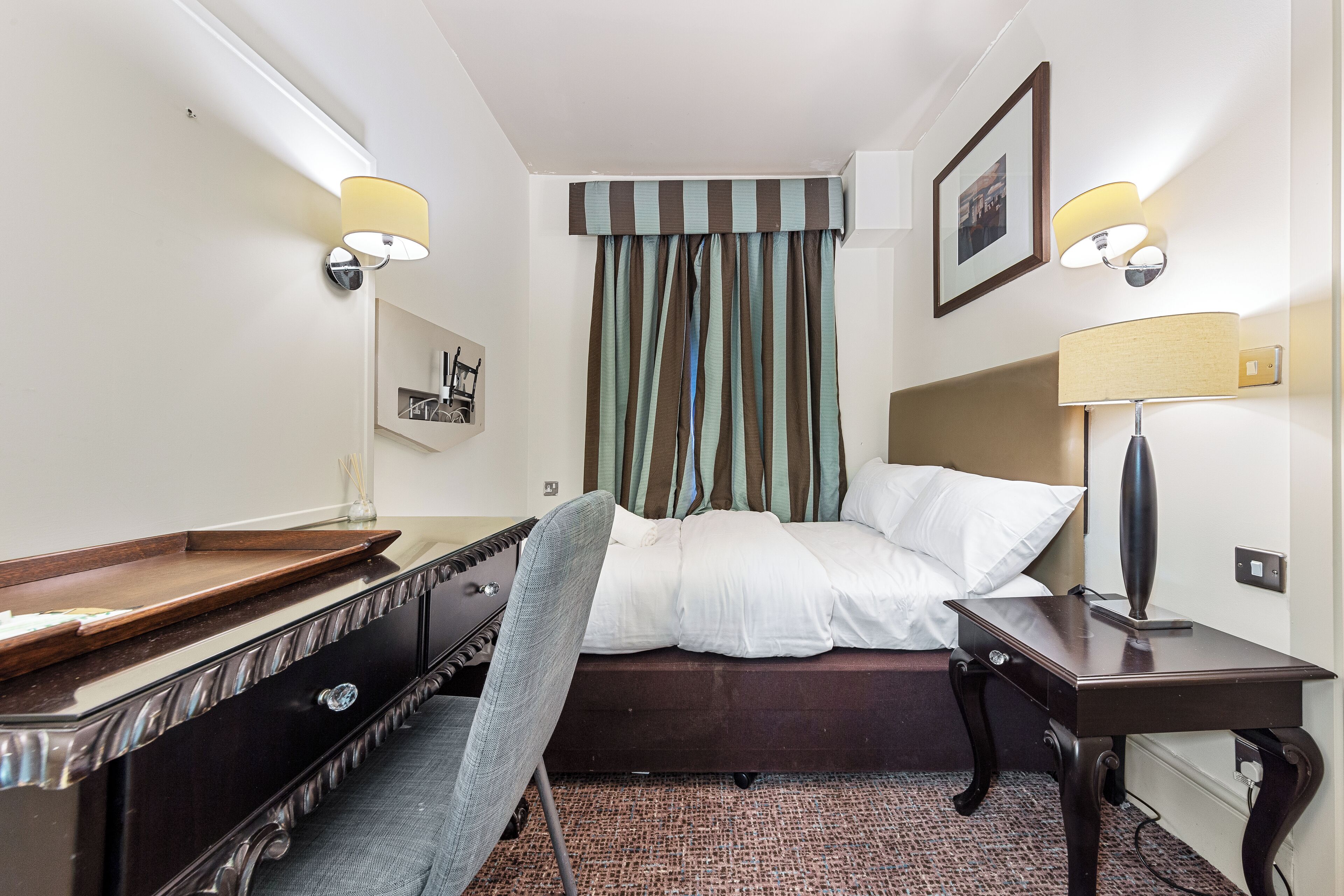 A hotel room with a bed, a desk, a chair, a lamp, and a picture on the wall.