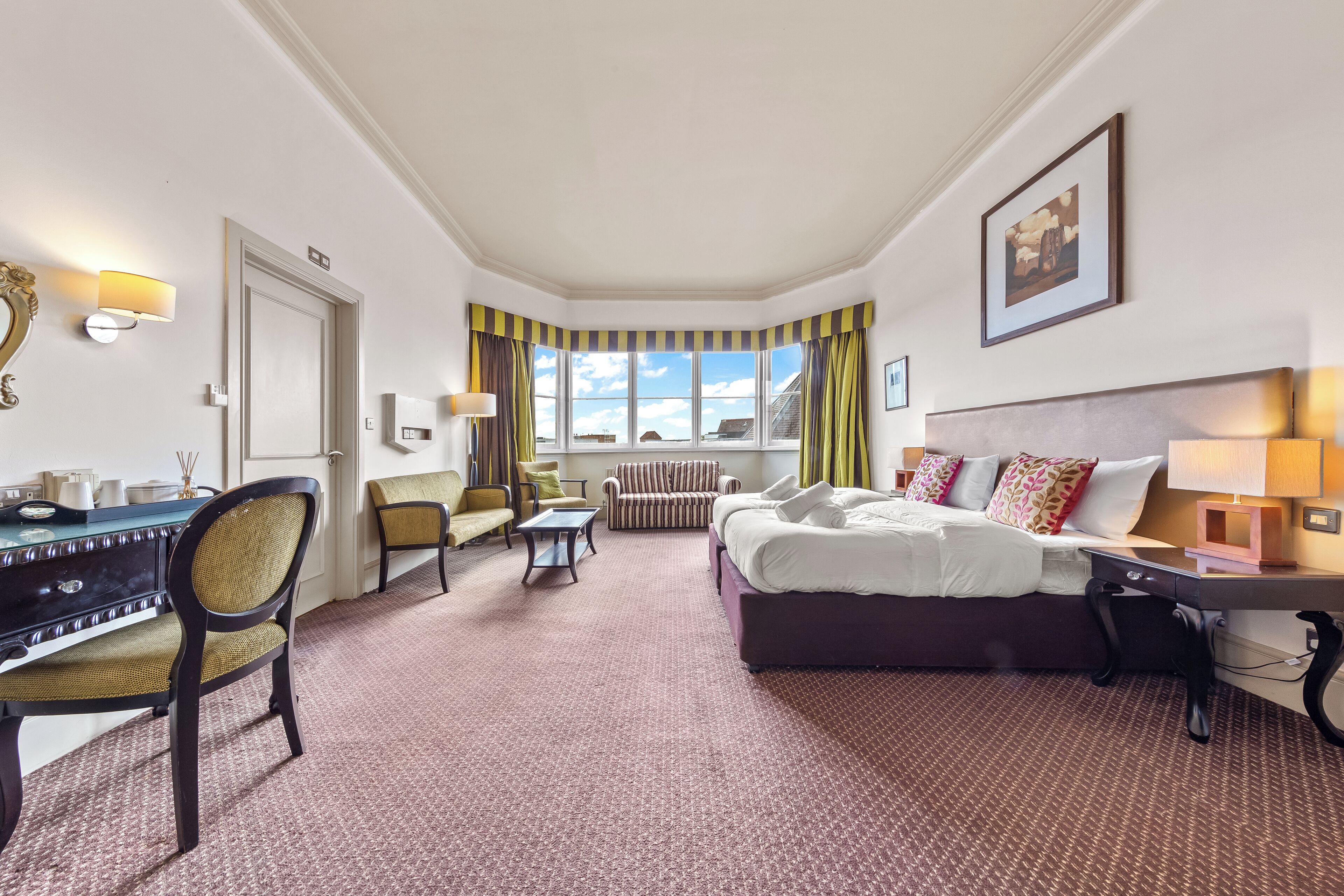 A hotel room with a bed, desk, chair, and a view of the sky through large windows.