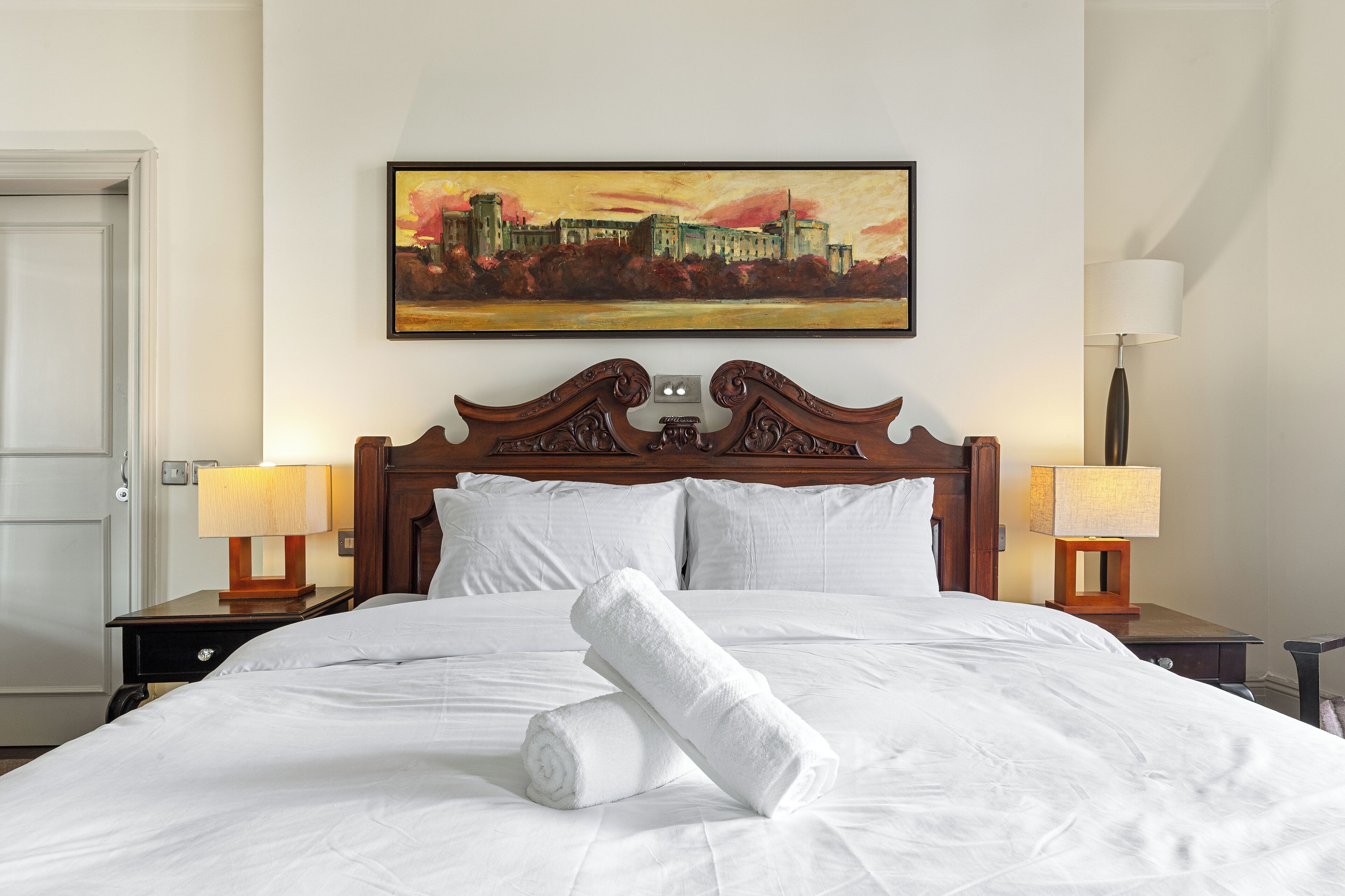 A neatly made bed with white linens, two bedside lamps, and a painting of a cityscape on the wall.