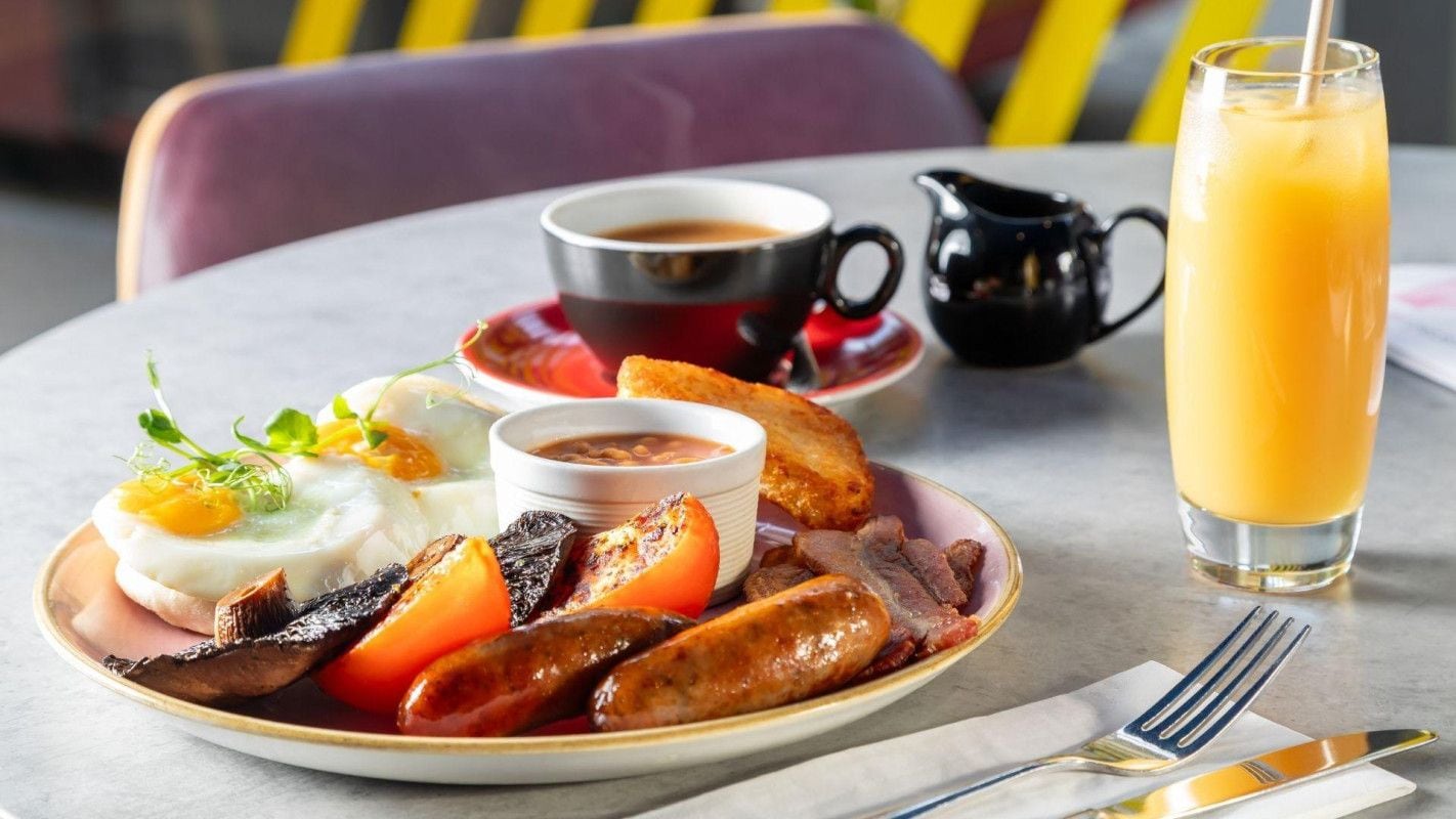 A breakfast plate with eggs, sausages, tomatoes, and beans, a cup of coffee, a pitcher, and a glass of orange juice on a table.
