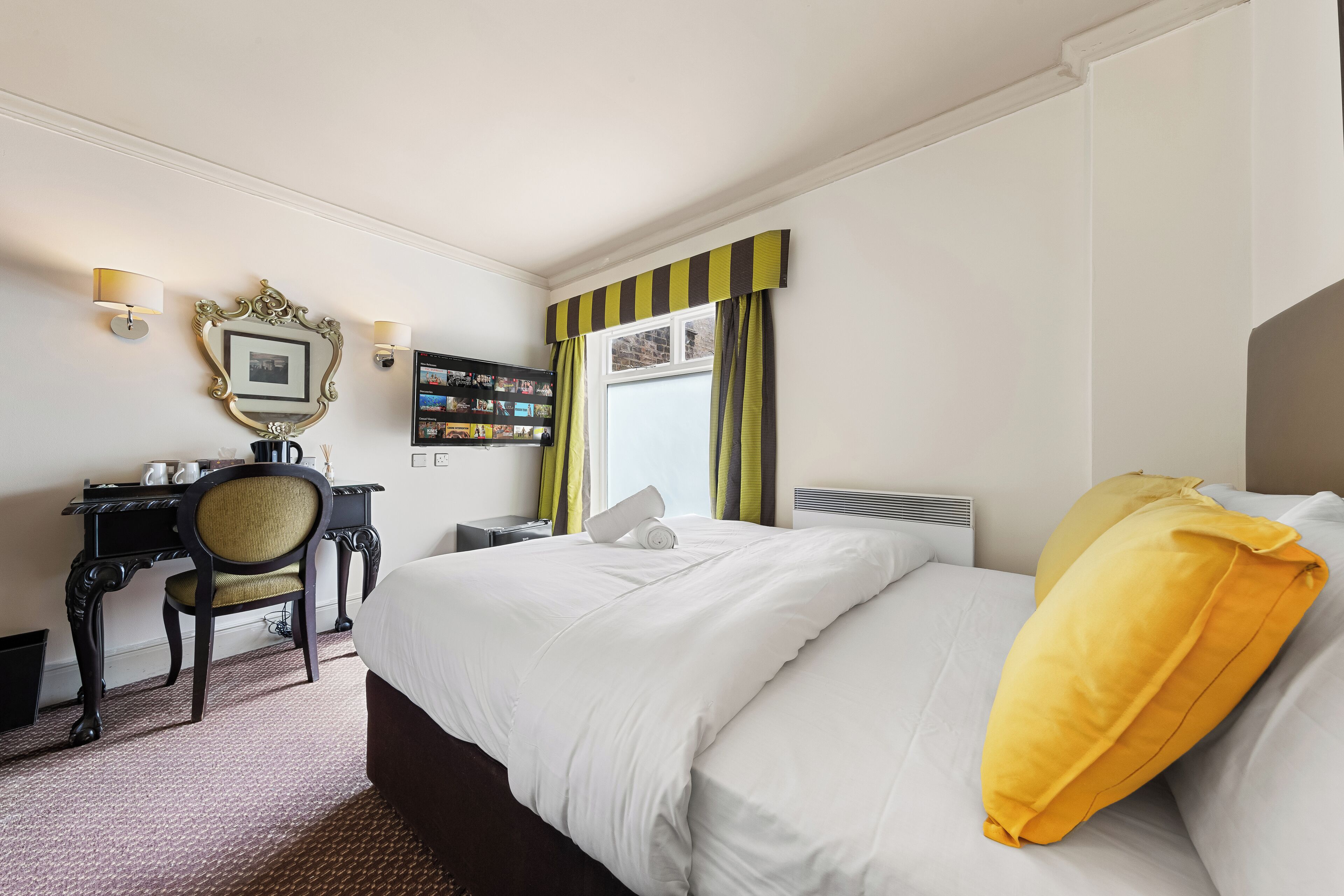 Hotellihuone, jossa on suuri sänky, työpöytä tuolilla, peili ja ikkuna, jossa on verhot.