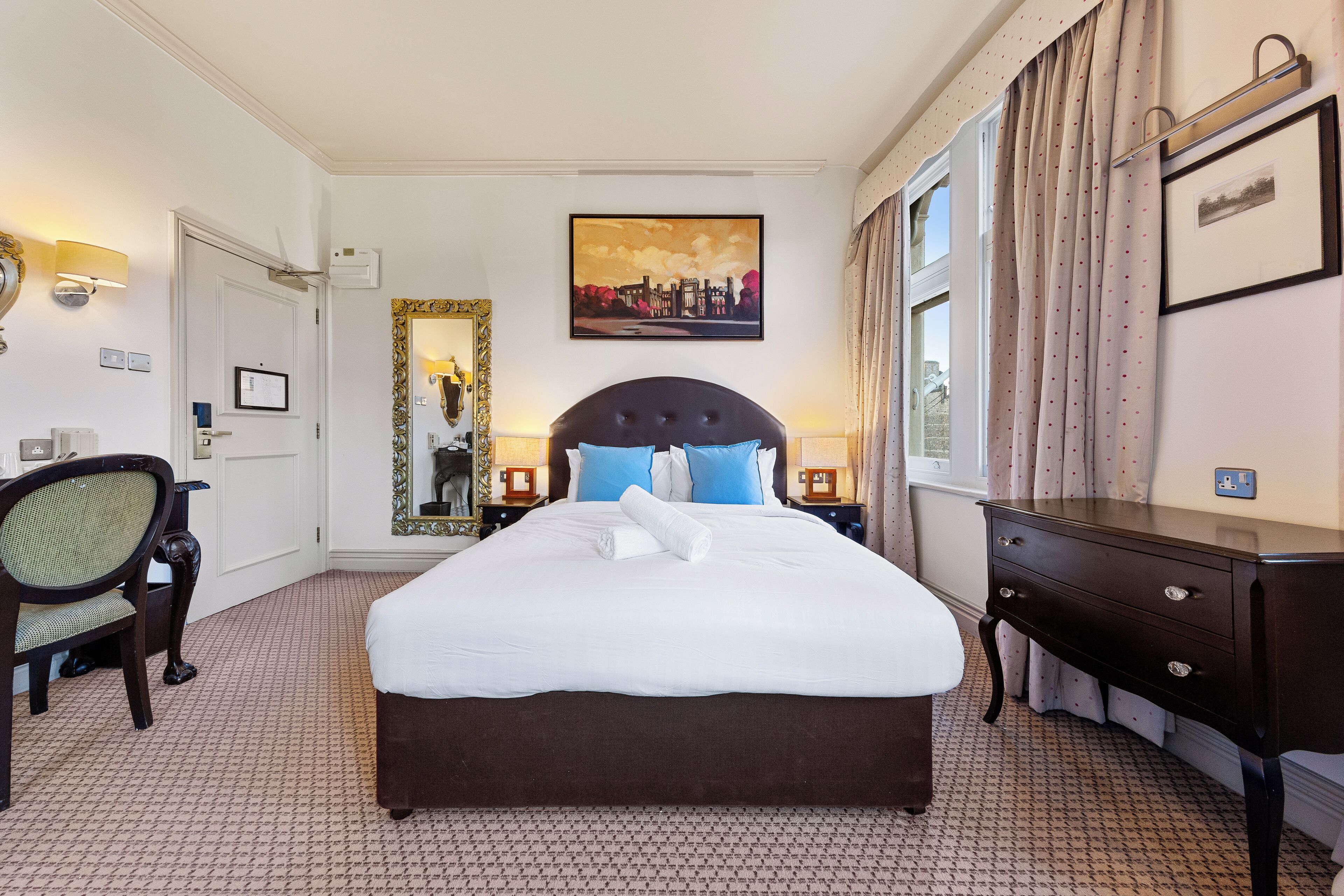 A neatly made bed with white linens and blue pillows, a wooden nightstand with a lamp, a chair, and a dresser with a mirror.