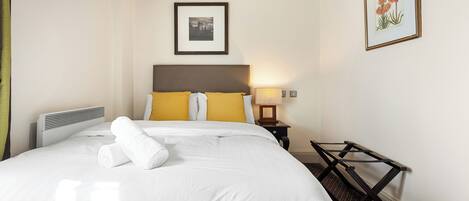 A neatly made bed with white linens and rolled towels, a bedside table with a lamp, a framed botanical print on the wall, and a small wooden stool.