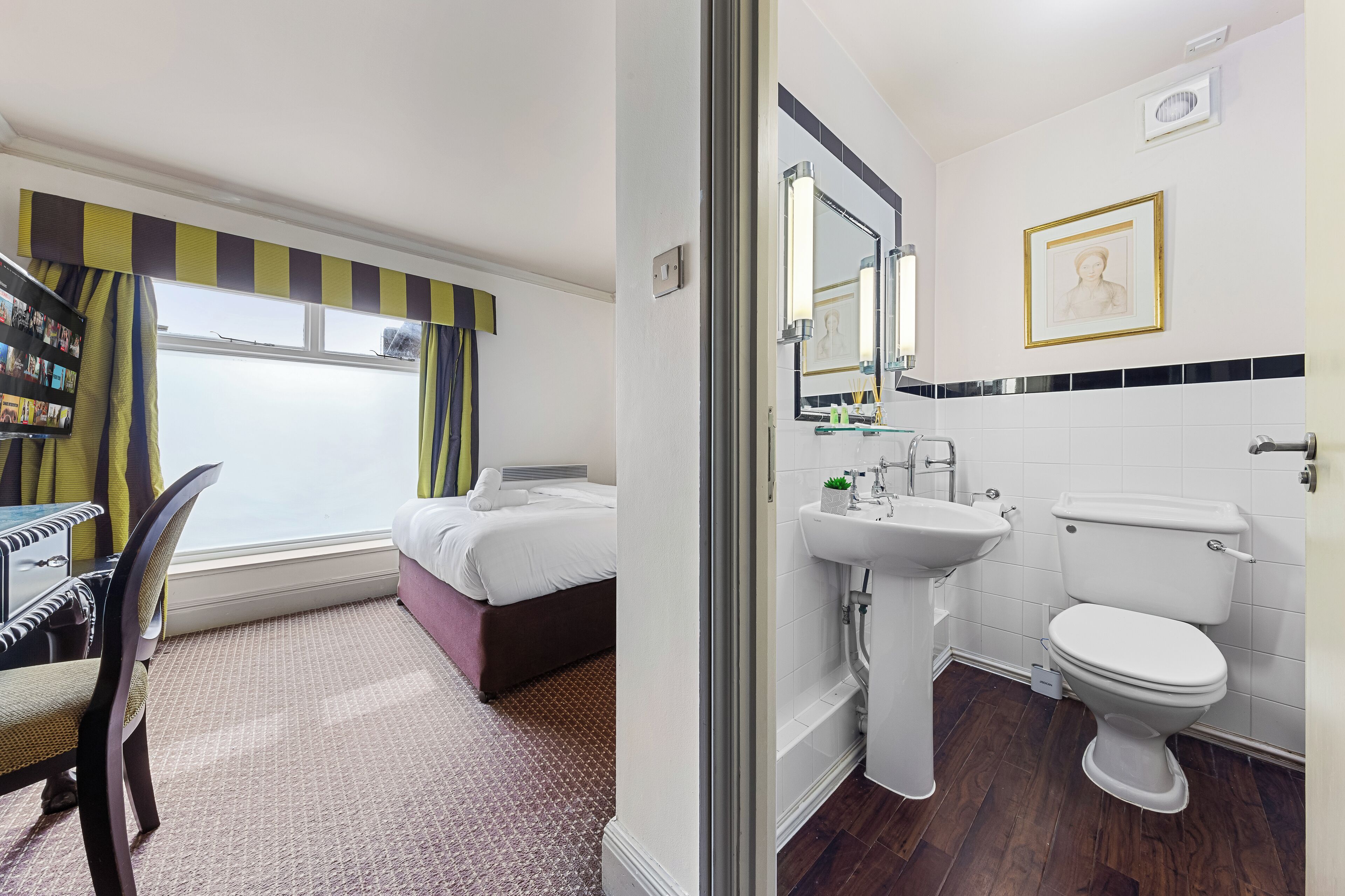 A hotel room with a bed, a desk, a chair, a window with curtains, and a bathroom with a sink, toilet, and mirror.