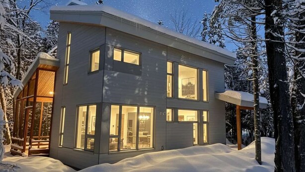 Una casa moderna de dos pisos con ventanas amplias y un porche cubierto, rodeada de árboles nevados.