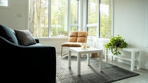 Un salón moderno con un sofá negro, un sillón marrón, una mesa de centro blanca y una mesita auxiliar pequeña. Amplias ventanas ofrecen vistas a un área boscosa.