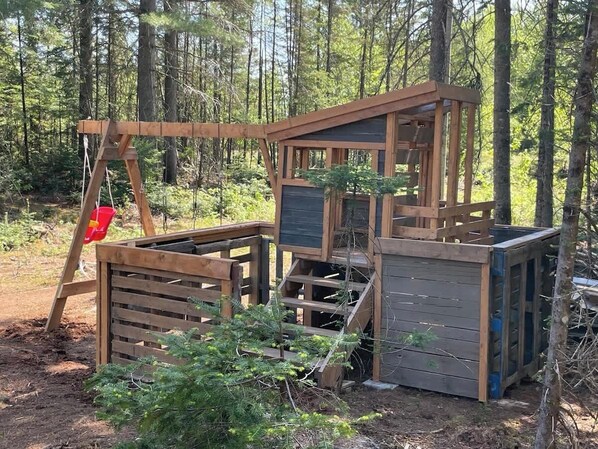 A wooden cabin structure in a forest setting with a ladder, a small enclosed area, and a platform.