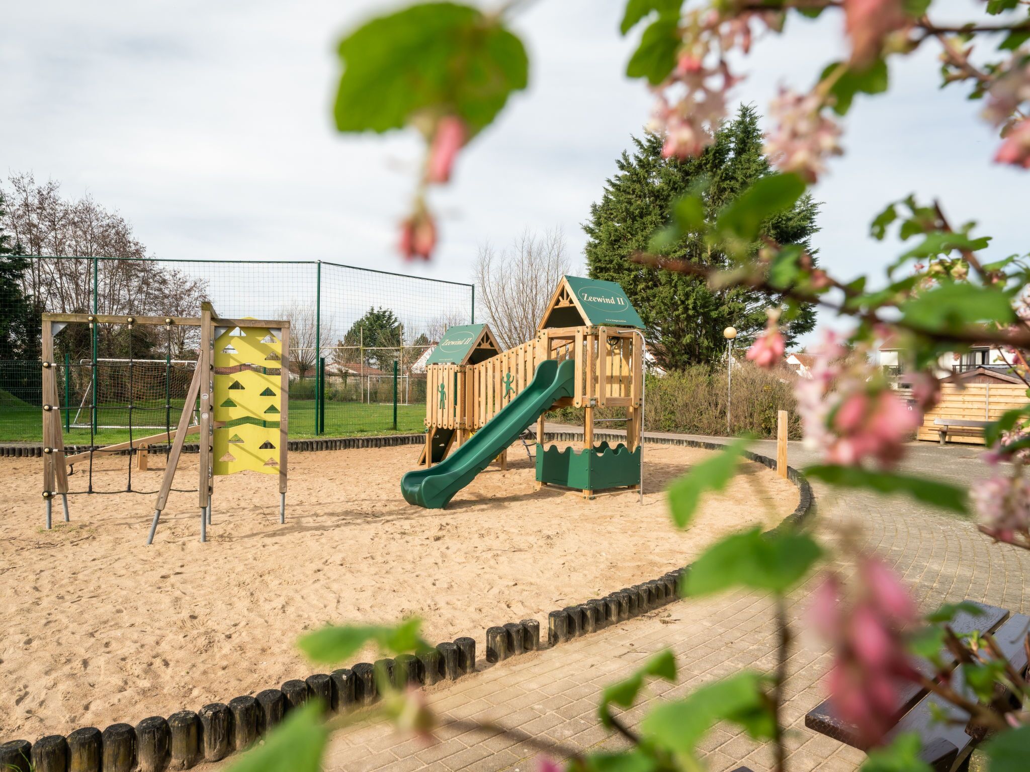 A playground with a slide and climbing structures, surrounded by a fence and sand area.