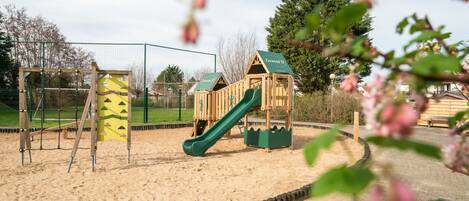 A playground with a slide and climbing structures, surrounded by a fence and sand area.