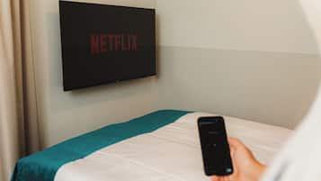 A hotel room with a bed, a wall-mounted TV displaying the Netflix logo, and a person holding a smartphone.