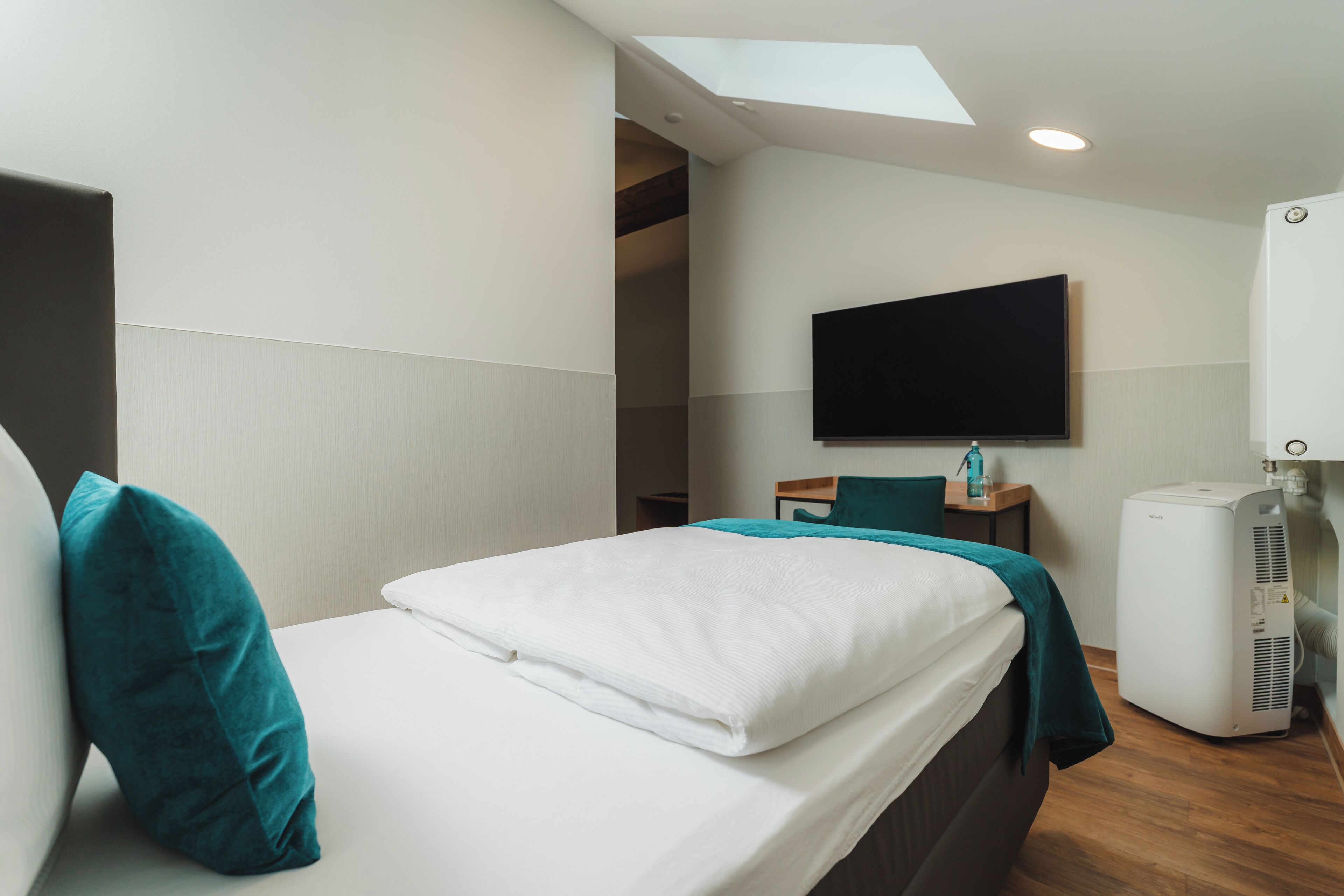 A modern hotel room with a bed, a flat-screen TV, a heater, and a skylight.