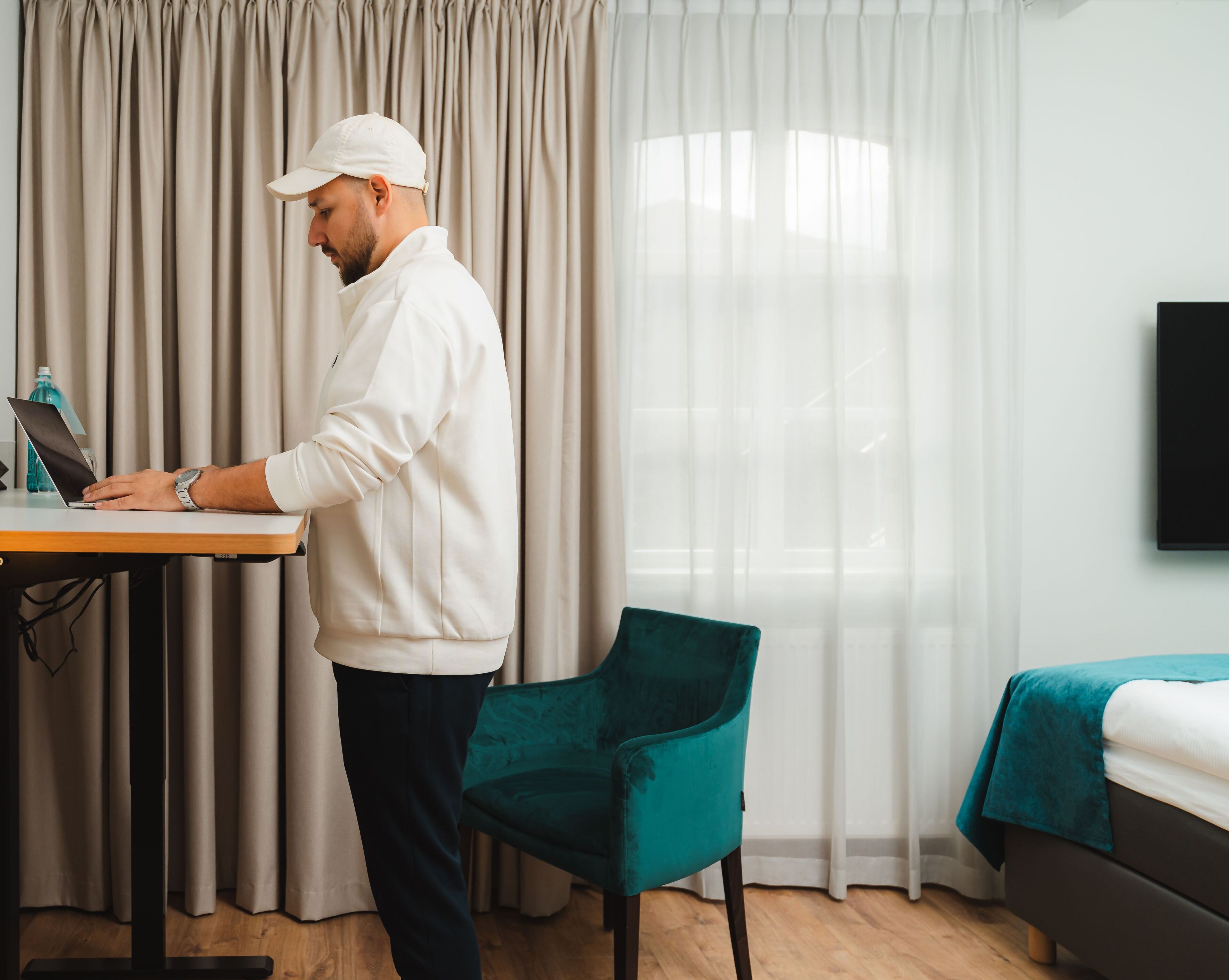 Ein Hotelzimmer mit einem Bett, einem Schreibtisch mit einem Laptop, einem türkisfarbenen Sessel und einem Fernseher an der Wand.