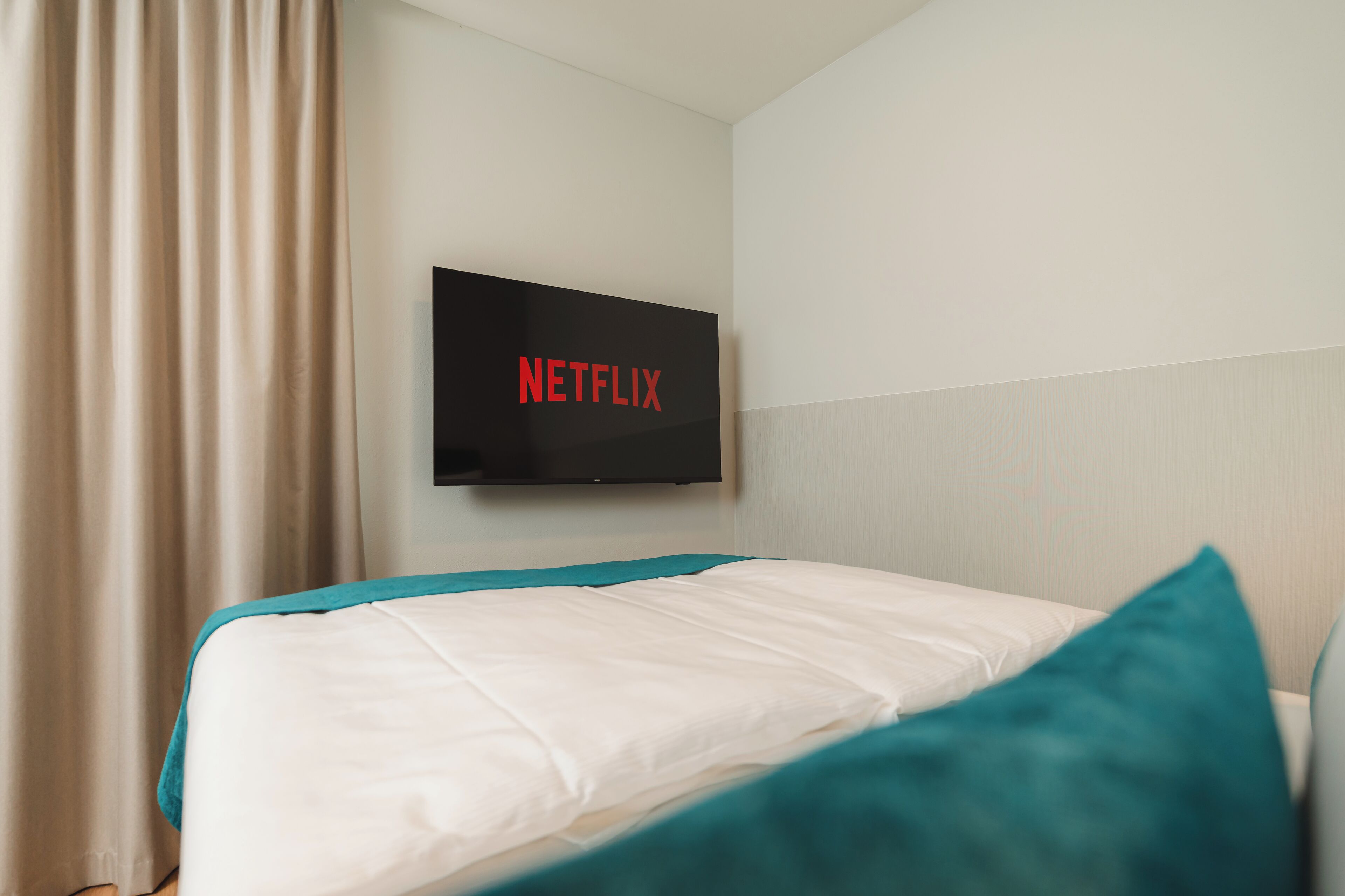 A hotel room with a bed, a TV mounted on the wall displaying the Netflix logo, and a window with curtains.