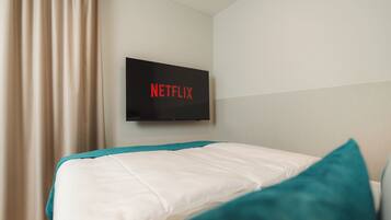 A hotel room with a bed, a TV mounted on the wall displaying the Netflix logo, and a window with curtains.