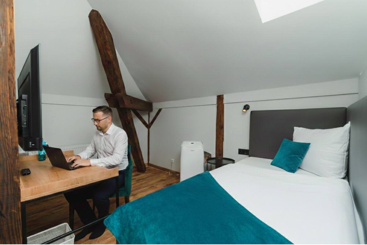Ein modernes Hotelzimmer mit einem Holztisch, einem Bett mit türkisfarbenem Bettzeug und einem großen Fenster.